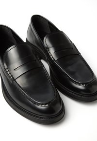 Schwarze Lederloafers mit glatter Oberfläche, ausgestattet mit einem gesteppten Design und einem erhöhten Lederstreifen an der Oberseite. Flache Sohle, schlanke Silhouette.