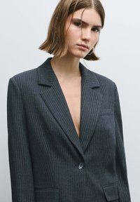 Donkergrijze blazer met krijtstreep, voorzien van ingelegde revers, één knoopsluiting en steekzakken aan de voorkant. Gestructureerde stof met een getailleerde pasvorm.