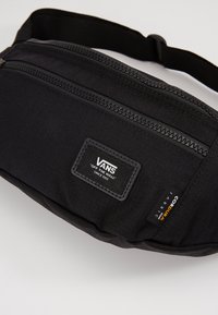Riñonera negra Vans hecha de tejido Cordura duradero. Cuenta con un bolsillo con cremallera en la parte frontal y una etiqueta tejida de la marca. Diseño compacto y práctico.