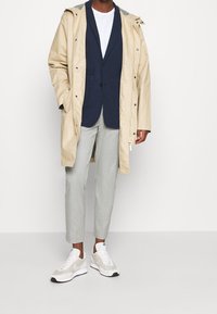 Beige raincoat with hood, navy blazer underneath, light gray trousers, and white sneakers. Textured fabric, tailored fit, and visible pockets.