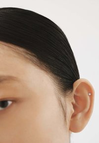 Primer plano de una oreja de tono de piel claro con un pequeño pendiente de plata, con el cabello oscuro y liso peinado hacia atrás contra un fondo neutro.
