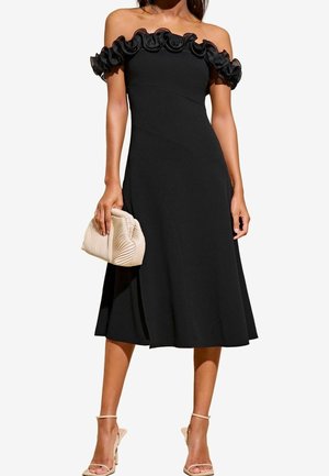 Nainen pukeutuneena mustaan olkapäät paljastavaan midimekkoon, jossa on röyhelöinen pääntie, pitää kädessään beigeä laskostettua clutch-laukkua ja käyttää beigen värisiä remmikenkiä.