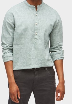 Homme portant une chemise vert clair à manches longues avec un col officier et des boutons, associée à un pantalon foncé, debout, les bras détendus.