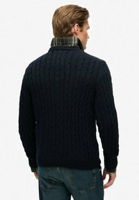 Mann von hinten gesehen, der einen navyblauen, grob gestrickten Pullover über einem grün-karrierten Hemd mit Kragen und blauen Jeans trägt, vor einem weißen Hintergrund.
