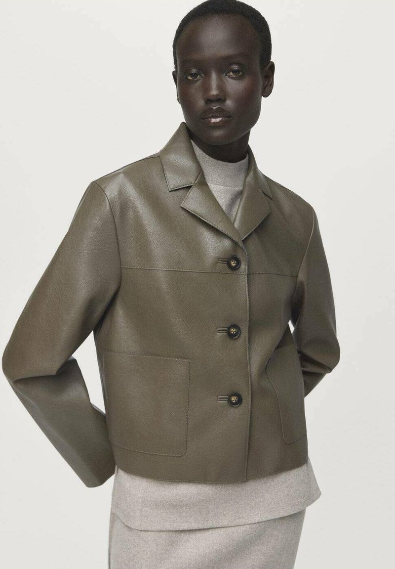 Personne à la peau foncée portant une veste en cuir vert olive à boutons sur un top en tricot gris clair et une jupe, se tenant devant un fond uni.