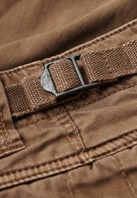 Bruine stof met een gestructureerde afwerking, voorzien van een metalen gesp en een geweven band, gestikt met contrasterende draden. Close-up van de materiaaldetails.