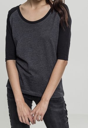 Femme portant un t-shirt à manches raglan gris foncé et noir avec un jean noir orné de détails lacés sur le côté, debout les mains jointes.