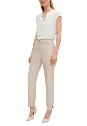 Femme portant un chemisier blanc sans manches, un pantalon beige ajusté avec des fentes aux chevilles, et des chaussures à talons hauts beiges assorties, debout sur un fond blanc.