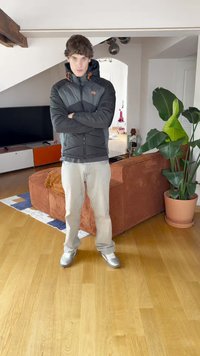Black insulated jacket with orange accents, light jeans, and silver sneakers. Indoor setting with a plant and a textured orange couch.