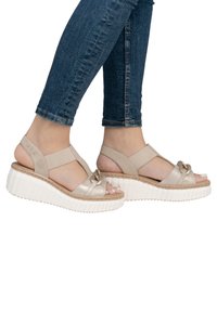 Sandales à plateforme beiges avec une semelle blanche texturée, dotées d'une sangle métallique et d'un nœud décoratif. Talon ouvert et sangles autour de la cheville pour un bon soutien.