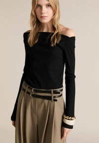 Haut noir à épaules dénudées en rib et à manches longues, associé à un pantalon beige taille haute avec des ceintures noires superposées et un bracelet accentué d'or.