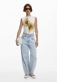 Fitted white sleeveless top with yellow floral design, paired with light blue baggy jeans featuring side button accents and white shoes.