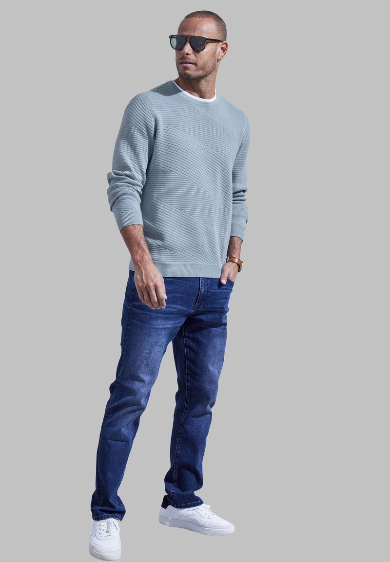 Helles blaues, strukturiertes Sweatshirt mit langen Ärmeln, kombiniert mit dunkelblauen Jeans und weißen Sneakers. Accessorized mit schwarzen Sonnenbrillen.