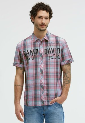 Man with curly hair wearing a short-sleeve red and blue plaid shirt with "Camp David" text and blue jeans, standing with one hand in pocket.