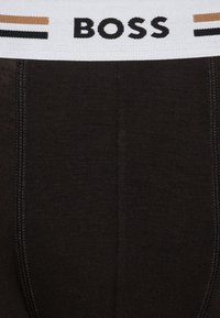 Čierne mužské boxerky s bielym pásom s logom "BOSS" v čiernej farbe, doplnené tenkými hnedými pruhmi. Vyrobené z hladkej, elastickej látky.