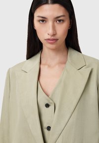 Blazer verde chiaro con una pattina strutturata e due bottoni neri, indossato sopra un top a scollo a V abbinato, realizzato in un tessuto liscio e testurizzato.