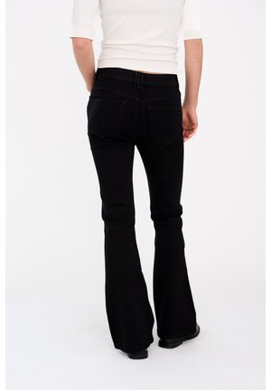 Persona che indossa jeans neri a zampa e un top bianco aderente, mostrata di spalle su uno sfondo bianco uniforme.