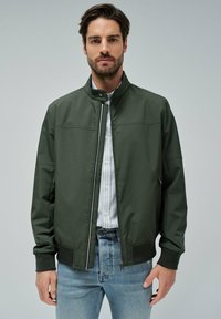 Chaqueta bomber de color verde oscuro con cuello alzado, cremallera frontal y puños elásticos. Combinada con una camisa clara de rayas y jeans azules.