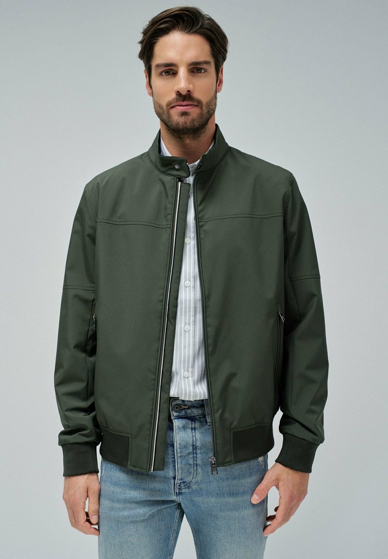 Chaqueta bomber de color verde oscuro con cuello alzado, cremallera frontal y puños elásticos. Combinada con una camisa clara de rayas y jeans azules.