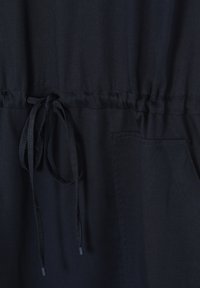 Tissu bleu foncé avec taille élastique froncée, cordon de serrage noué au centre et poche plaquée sur le côté droit.