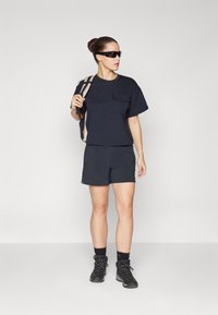 Camiseta de manga corta azul marino con un bolsillo, combinada con pantalones cortos negros. El modelo lleva gafas de sol, calcetines oscuros y botas de senderismo negras. Mochila colgando del hombro.