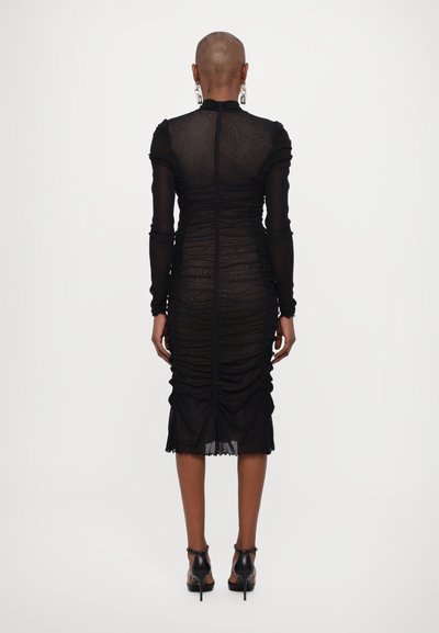 Rochie neagră cu pliuri, cu mâneci lungi din material transparent, o siluetă aderentă și o lungime până la jumătatea gambei. Închidere cu un fermoar la spate. Tocuri negre.