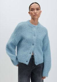 Donna che indossa un cardigan buttoned di un blu chiaro e jeans grigio scuro, in piedi contro uno sfondo chiaro e uniforme.