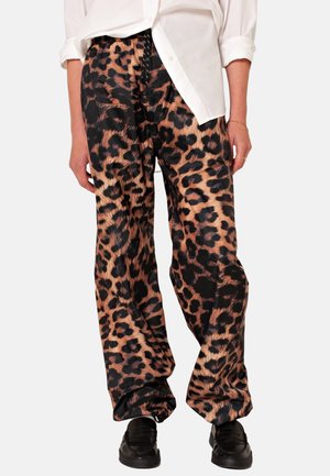 Persoon draagt losse broek met luipaardprint, wit overhemd met opgerolde mouwen en zwarte schoenen, staande tegen een effen achtergrond.