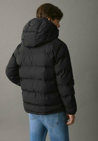 Svart puffjacka med huva, med en kviltad design och elastiska ärmar. Buren över blå jeans, sedd bakifrån.