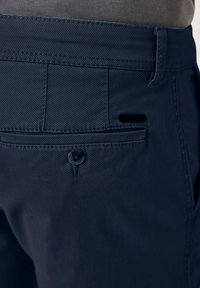 Marineblaue Hose aus strukturiertem Stoff, mit einer Gesäßtasche und Knopfverschluss sowie zusätzlichem seitlichen Eingriff. Sichtbare Steppnähte.