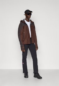 Bruine en grijze windbreakerjas met een ritssluiting aan de voorkant en een capuchon, gecombineerd met zwarte broeken en schoenen. Accessoires omvatten een zwarte pet en een zonnebril.