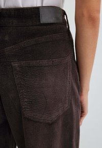 Pantaloni di velluto a coste marroni con finitura testurizzata, dotati di una tasca posteriore e di una toppa con logo in pelle sulla cintura, che evidenziano il motivo rigato del tessuto.