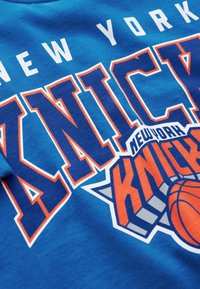 Kék pamut póló, amelyen "NEW YORK KNICKS" felirat található narancssárga és fehér színben, kosárlabda grafikával és csillag díszítéssel.
