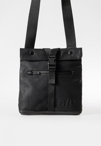 Black bag made of a matte fabric. Features a flat top, adjustable strap, zippered pocket, and embossed logo. Minimalist design.