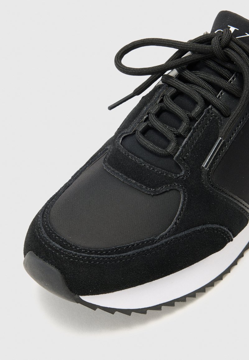 Chaussure de sport noire dotée d'un dessus en synthétique lisse et d'accents en daim, avec un bout rond, une semelle texturée et des lacets plats.