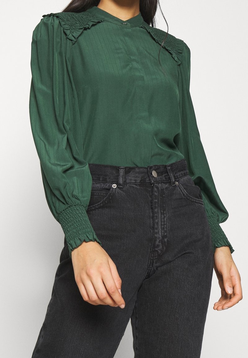 Femme portant un chemisier vert foncé à manches longues avec épaules et poignets froncés, rentré dans un jean noir taille haute.