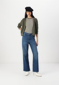 Zelený pletený cardigan cez čiernobielo pruhované tričko, skombinovaný s voľnými modrými džínsami a bielymi čižmami na podpätku. Čierny baret na hlave.