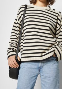 Pull en tricot rayé crème et noir avec un col et des poignets côtelés. Présente des détails de bouton sur l'épaule. Associé à un jean bleu clair.