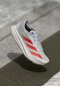 Lichtgrijze sportschoen met rode accenten, bovenkant van mesh, vetersysteem en rubberen zool. Bevat drie strepen branding.