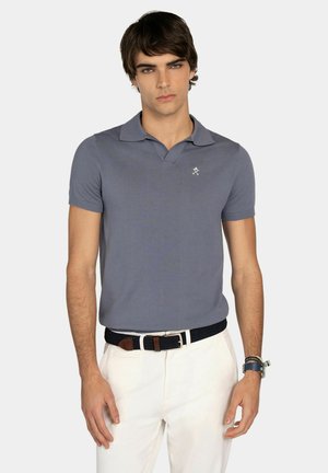 Joven vestido con una camisa polo de manga corta gris, pantalones blancos, cinturón trenzado negro y reloj de pulsera, posando frente a un fondo liso.