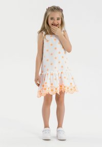 Vit klänning med orange blommönsterbroderi, scallopad nederkant och volang längst ner. Modell som bär vita sneakers, står med handen på munnen.