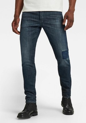 Dunkelblaue Jeans aus Denim mit schmaler Passform, versehen mit einem abgenutzten Fleck am rechten Knie, kombiniert mit schwarzen Stiefeletten.