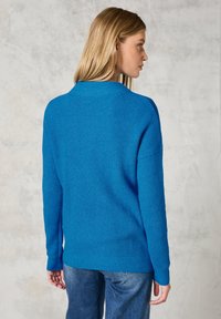 Blauer gestrickter Pullover mit einem strukturierten Muster, gerippten Bündchen und entspannter Passform, von hinten vor einem neutralen Hintergrund gezeigt.