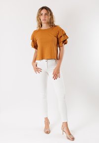 Top corto a maniche corte color senape con maniche arricciate, abbinato a jeans skinny bianchi e sandali con cinturino dorati. Sfondo semplice.