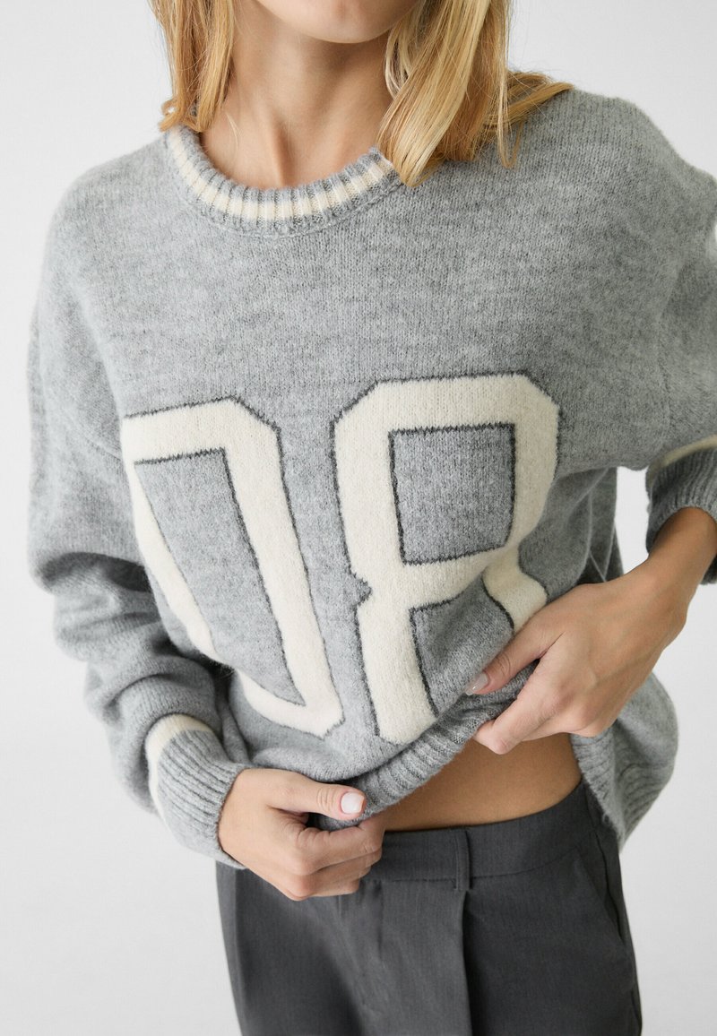 Grauer Strickpullover mit einem auffälligen cremefarbenen "08"-Design, geripptem Kragen und Bündchen sowie einer weichen Textur. Getragen mit grauen Hosen.