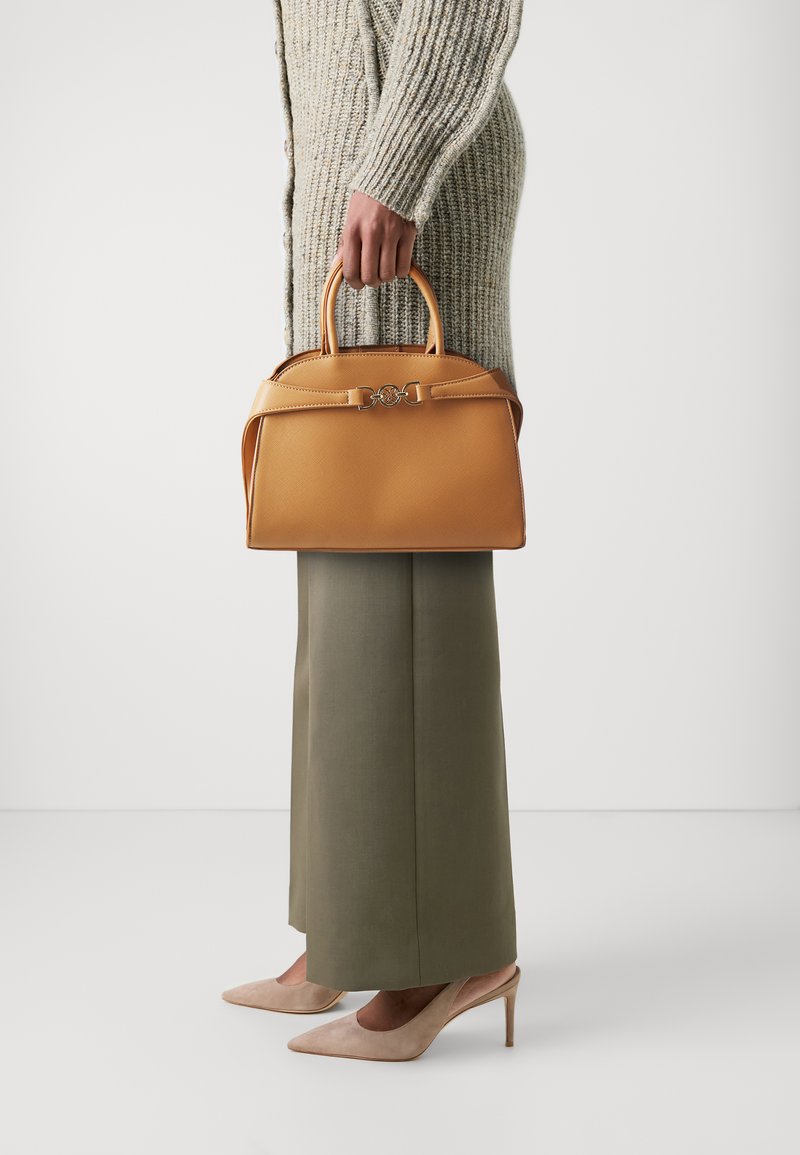 Bolso de cuero marrón claro con un diseño estructurado, asas superiores y un detalle decorativo en la correa. Lleva puesto un cárdigan de punto y pantalones verdes.