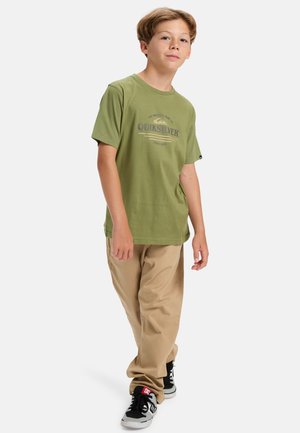 Camiseta de algodón verde con el logo de "Quiksilver" en azul, combinada con pantalones beige y zapatillas negras con detalles en blanco. Pose de pie.