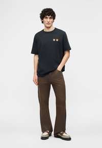 Bărbat cu păr creț, purtând un tricou negru cu două mici broșe în formă de animal, pantaloni maro și adidași bej, stând în fața unui fundal simplu.