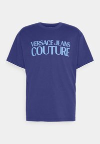 Camiseta de algodón azul con mangas cortas y cuello redondo, que presenta un gran texto en blanco que dice "VERSACE JEANS COUTURE" en la parte delantera.