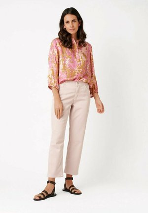 Donna in piedi che indossa una camicetta floreale rosa e oro, pantaloni beige chiaro e sandali neri con cinturini su sfondo bianco.